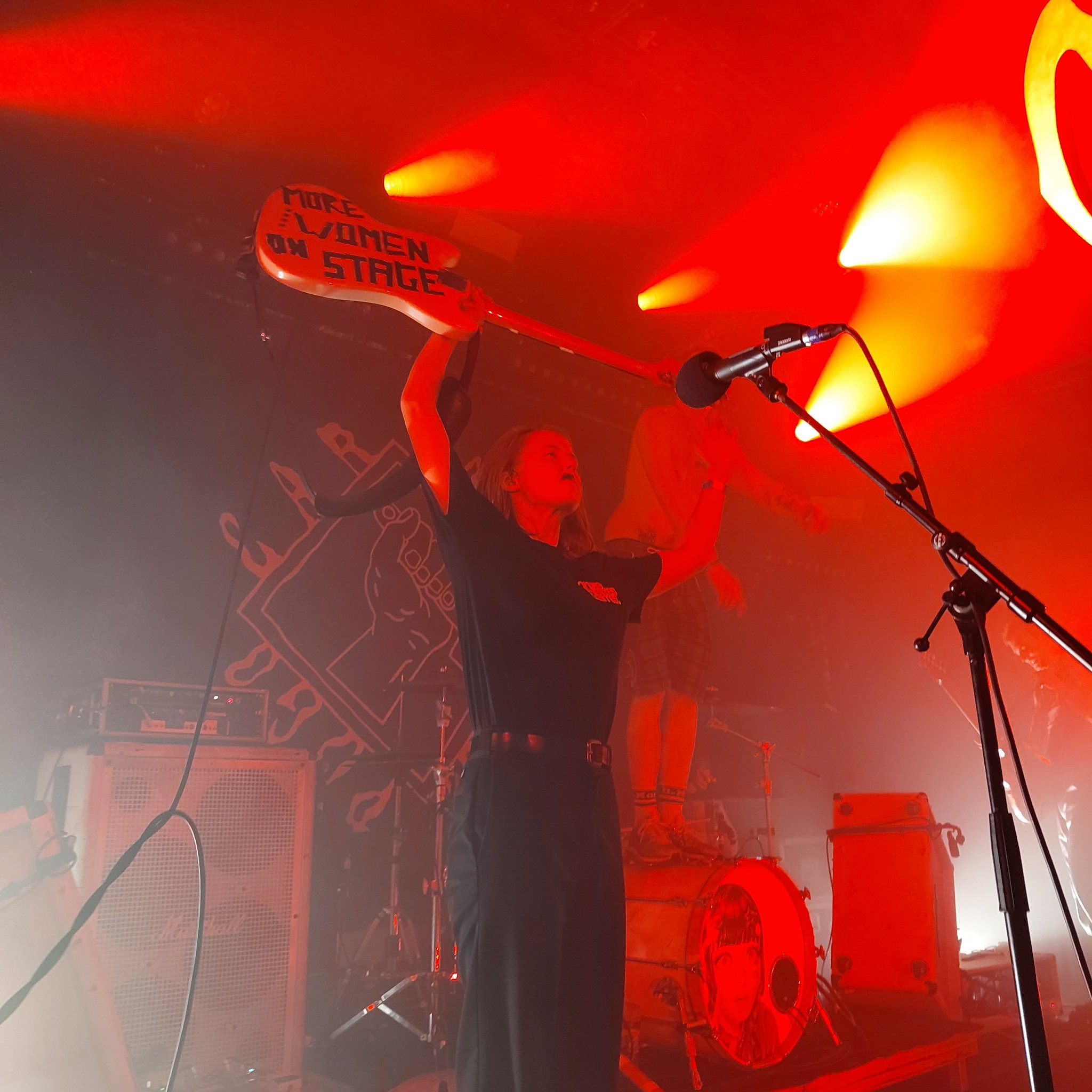 Photo de Lola Frichet qui montre sa basse ou est écrit au gaffer « MORE WOMEN ON STAGE »