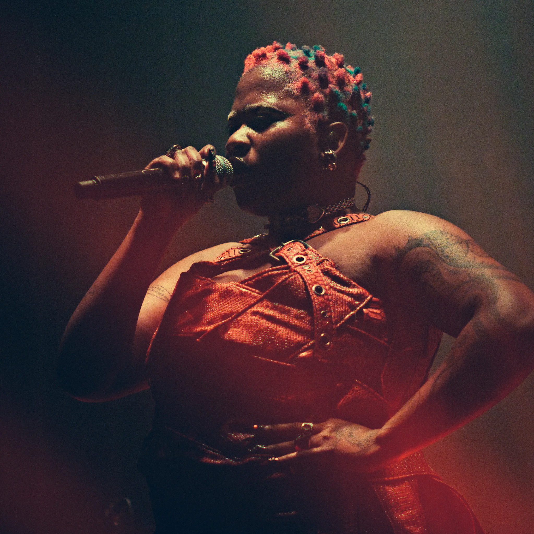 Photo d’Uzi en train de chanter, sa main gauche sur sa hanche et sa main droite tenant le micro devant sa bouche.
Elle à les cheveux court, teint en rouge et bleu.