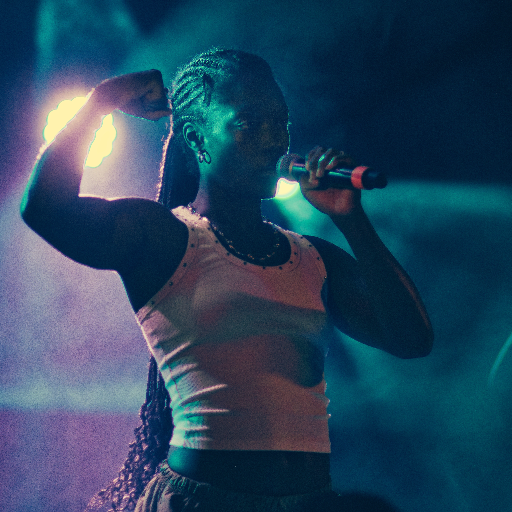 Photo de Shani montant son biceps droit gainé, sa main gauche tien son micro.
La lumière est composée de bleu et de violet.
