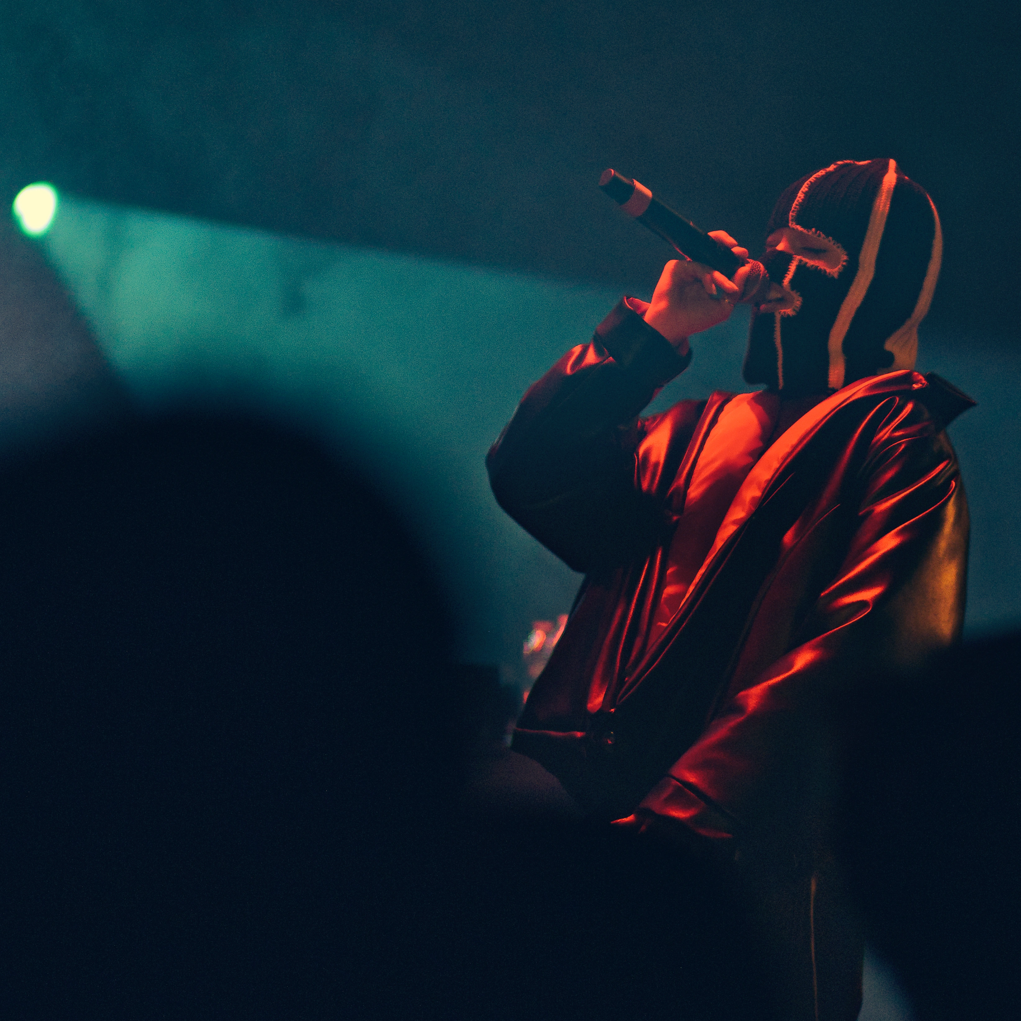 Chanteuse cagoulée avec un gros manteau, éclairée en rouge dans une scène de brume bleu.
Elle tien son mico de la main droite.