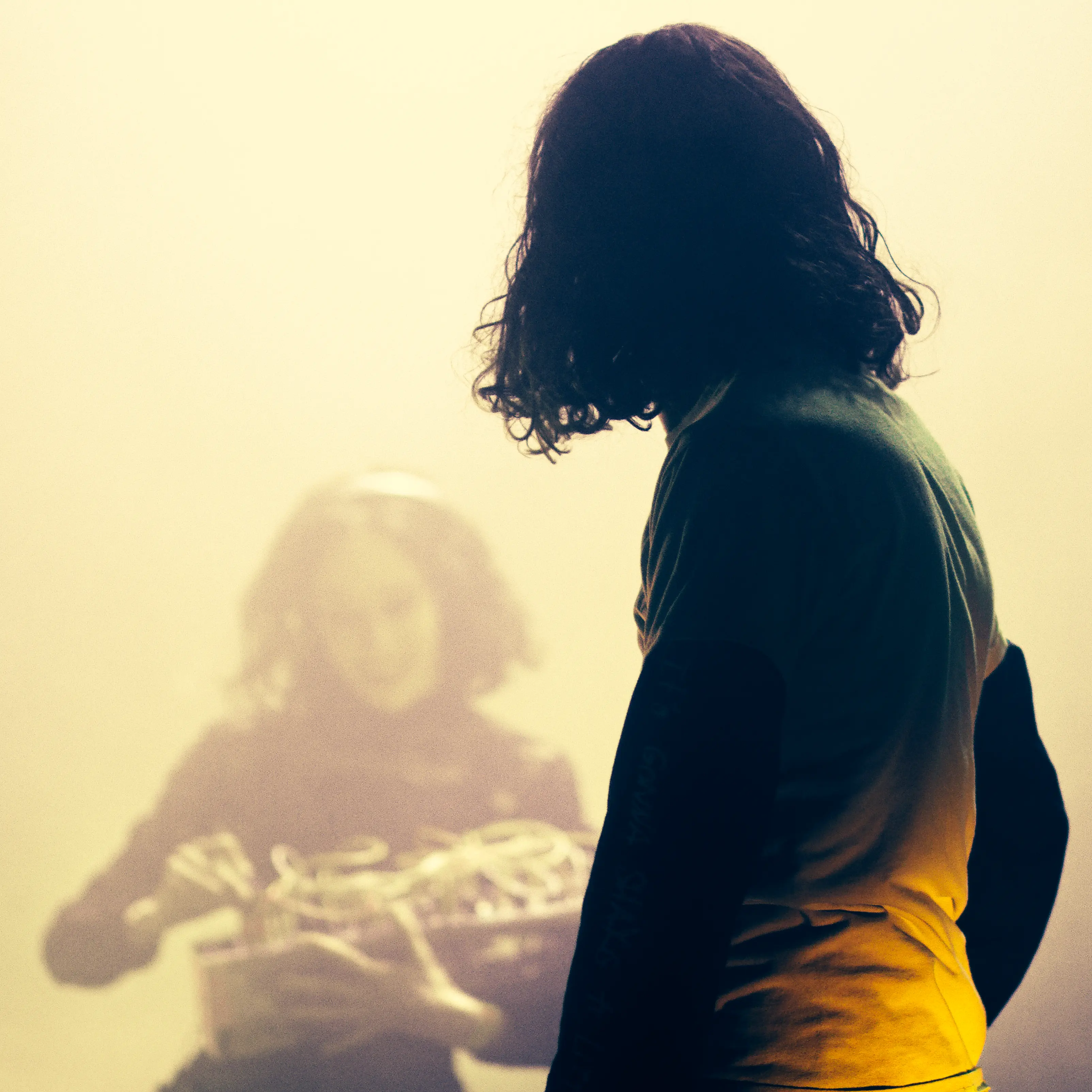 Photo mise au point sur le musicien de dos qui regarde la musicienne, en flou dans la fumée.
Elle tien un synthétiseur avec beaucoup de cables dans ses bras