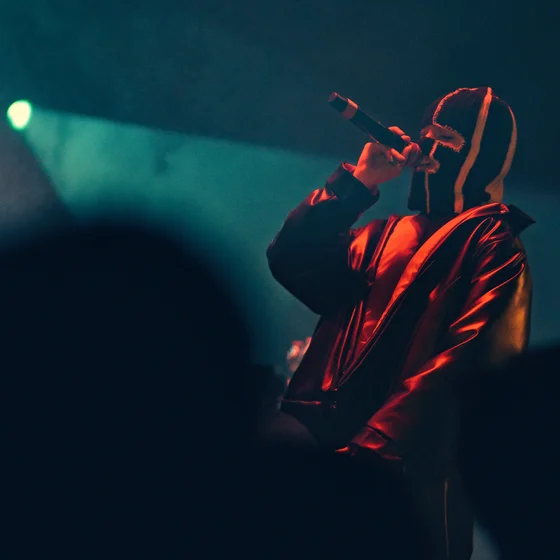 Chanteuse cagoulée avec un gros manteau, éclairée en rouge dans une scène de brume bleu.
Elle tien son mico de la main droite.