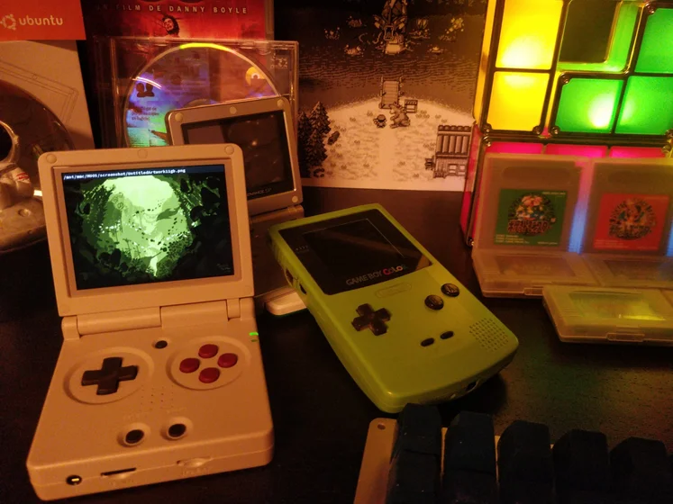 Photo d’un étage d’étagère avec une gameboy color verte, une gbSP grise, des cartouches japonaise de pokémon vert et rouge…
Le sujet de la photo est une console d’émulation qui affiche un pixel art réalisé par Menica