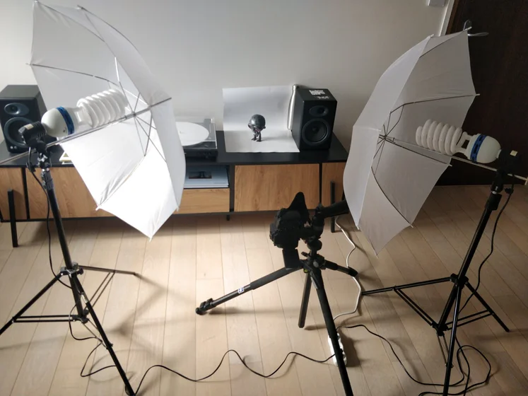 Studio photo improvisé sur un meuble de salon.
Un trépied photo et des lumières sur pied sont installés pour photographier une figurine posée sur une grande  feuille blanche.