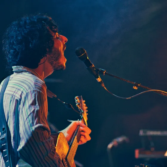 Photo du chanteur en train de jouer de la guitare, bouche ouverte.
Éclairage d’ambiance bleu, une lumière rouge sur son visage